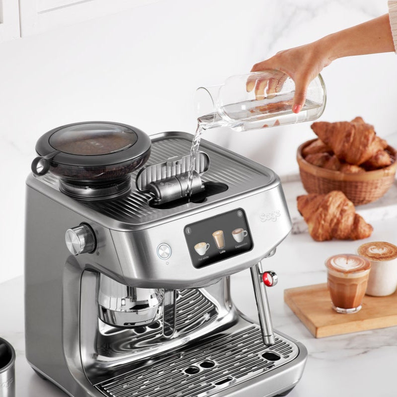 Coffee machine with a person pouring liquid into it, surrounded by croissants and coffee cups on a white surface.