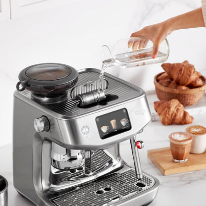 Coffee machine with a person pouring liquid into it, surrounded by croissants and coffee cups on a white surface.